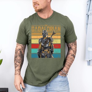 K&ouml;nnte beinhalten: Olivgr&uuml;nes T-Shirt mit einer Grafik des Mandalorian-Charakters und dem Text "The Mandalorian". Das Design zeigt eine Figur mit Helm und Speer vor einem gestreiften Hintergrund in Gelb, Orange, Rot und Blau.