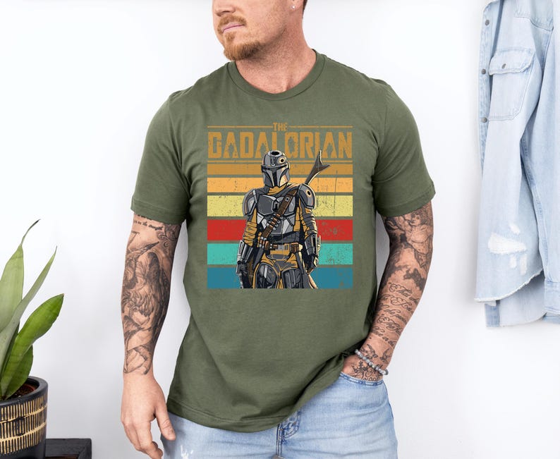 K&ouml;nnte beinhalten: Olivgr&uuml;nes T-Shirt mit einer Grafik des Mandalorian-Charakters und dem Text "The Mandalorian". Das Design zeigt eine Figur mit Helm und Speer vor einem gestreiften Hintergrund in Gelb, Orange, Rot und Blau.