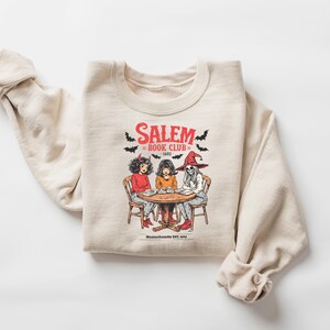 Puede incluir: Sudadera beige con un gr&aacute;fico de tres figuras sentadas alrededor de una mesa, con el texto "Salem Book Club 1692" y murci&eacute;lagos. Las figuras incluyen una mujer con cuernos de diablo, una mujer y un esqueleto con un sombrero de bruja.