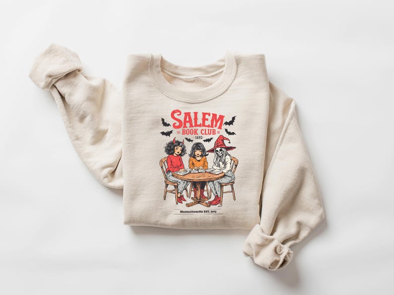 Puede incluir: Sudadera beige con un gr&aacute;fico de tres figuras sentadas alrededor de una mesa, con el texto "Salem Book Club 1692" y murci&eacute;lagos. Las figuras incluyen una mujer con cuernos de diablo, una mujer y un esqueleto con un sombrero de bruja.
