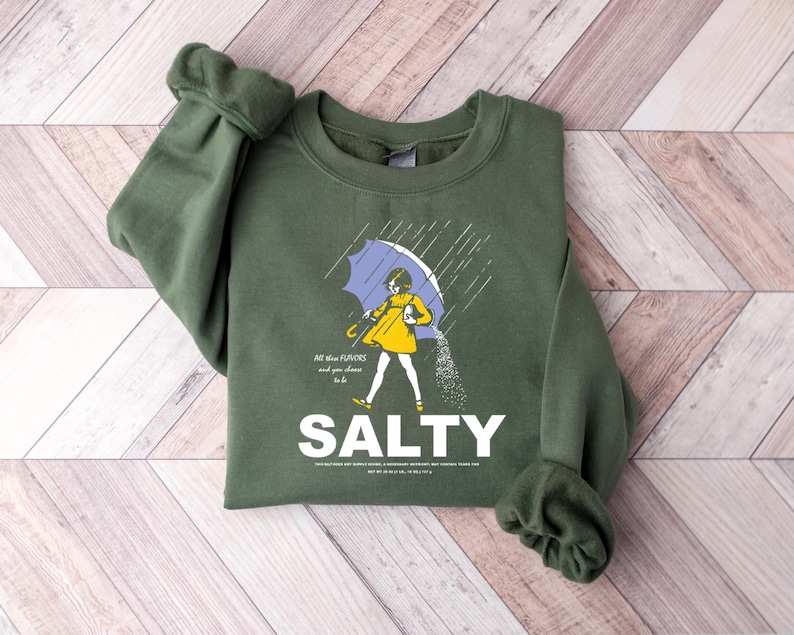 May include: An olive green sweatshirt with a graphic of a person under a blue umbrella, with the word "SALTY" in white. The person is wearing a yellow dress and is sprinkling salt. The text "All these flavors and you choose to be" is also present.
