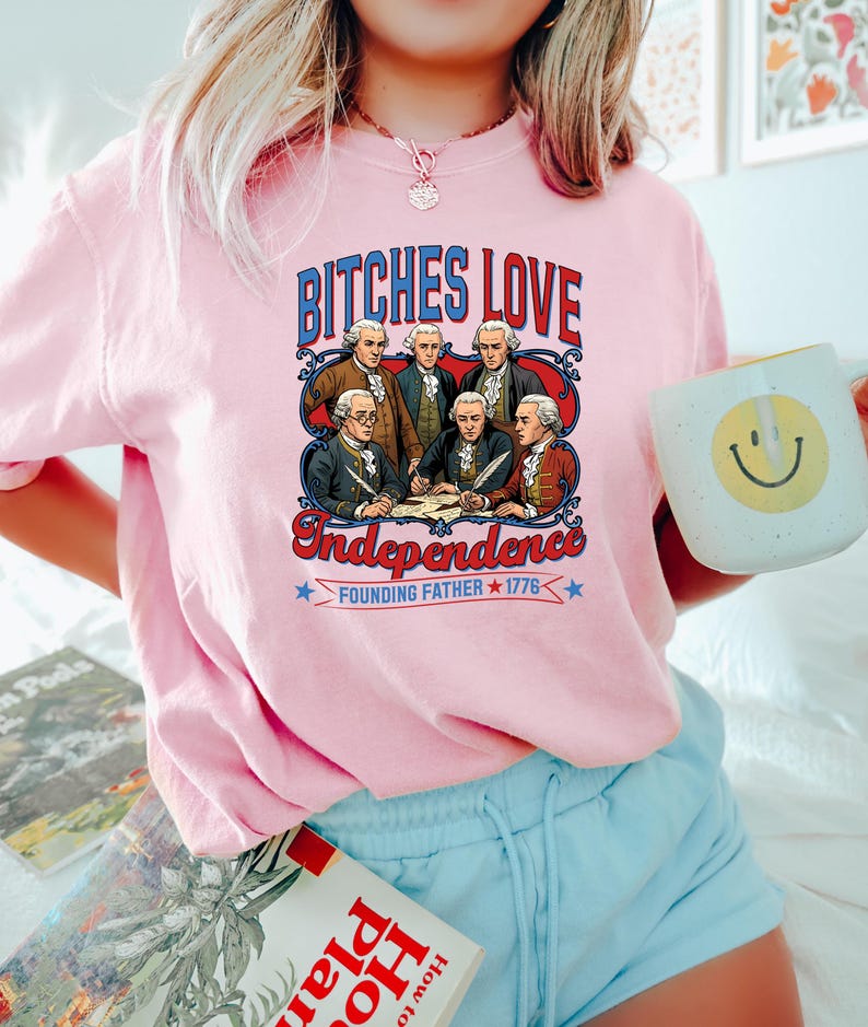 Może przedstawiać: R&oacute;żowa koszulka z napisem "BITCHES LOVE Independence" nad wizerunkiem Ojc&oacute;w Założycieli. Koszulka ma r&oacute;wnież napis "FOUNDING FATHER 1776" poniżej. W dłoni trzymany jest biały kubek z ż&oacute;łtą buźką.