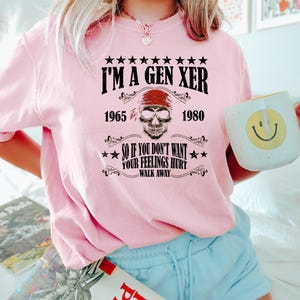 Op de afbeelding: Roze T-shirt met een zwarte afbeelding met de tekst "I'm a Gen Xer 1965 - 1980" en "So if you don't want your feelings hurt - walk away". De afbeelding bevat ook een schedel met een rode bandana.