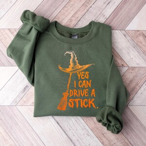 May include: An olive green sweatshirt with the text "YES I CAN DRIVE A STICK." The design features an orange witch hat and broom. The sweatshirt is folded on a light wood-grain surface.