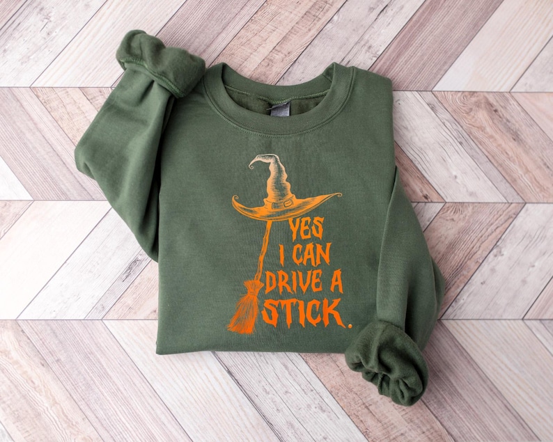 May include: An olive green sweatshirt with the text "YES I CAN DRIVE A STICK." The design features an orange witch hat and broom. The sweatshirt is folded on a light wood-grain surface.