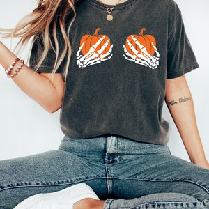 May include: Dark grey t-shirt with a Halloween design. The graphic depicts two orange pumpkins held by white skeleton hands. The shirt is worn with blue jeans and white slip-on shoes, creating a casual, seasonal style.