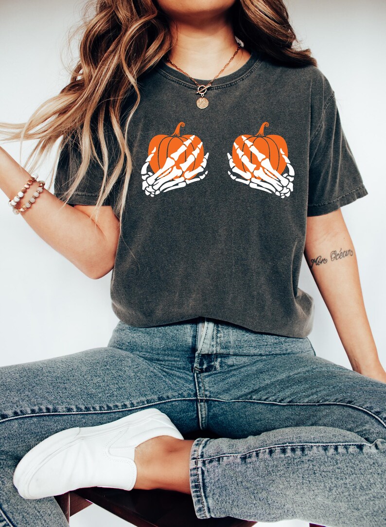 May include: Dark grey t-shirt with a Halloween design. The graphic depicts two orange pumpkins held by white skeleton hands. The shirt is worn with blue jeans and white slip-on shoes, creating a casual, seasonal style.