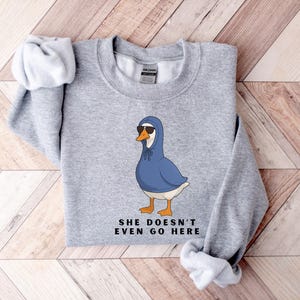 May include: A light grey sweatshirt with a cartoon goose wearing sunglasses and a blue hoodie. The goose has orange feet and a white belly. The text "SHE DOESN'T EVEN GO HERE" is printed below the goose in black.