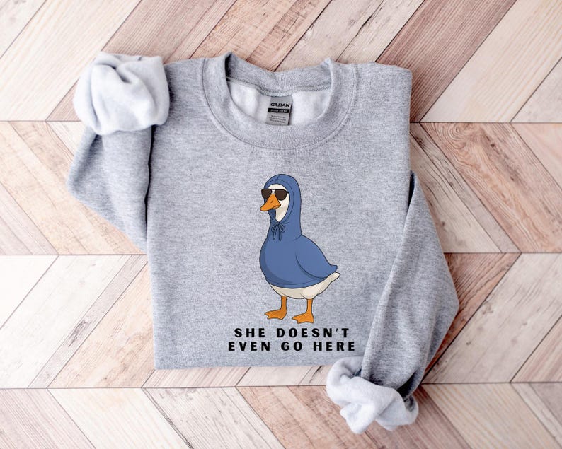 May include: A light grey sweatshirt with a cartoon goose wearing sunglasses and a blue hoodie. The goose has orange feet and a white belly. The text "SHE DOESN'T EVEN GO HERE" is printed below the goose in black.