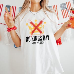 May include: White t-shirt with a graphic that says "NO KINGS DAY" with a crossed-out crown design. The date "JUNE 14th, 2025" is printed below the text. The background includes American flags and red, white, and blue decorations.