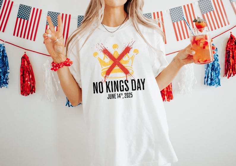 May include: White t-shirt with a graphic that says "NO KINGS DAY" with a crossed-out crown design. The date "JUNE 14th, 2025" is printed below the text. The background includes American flags and red, white, and blue decorations.