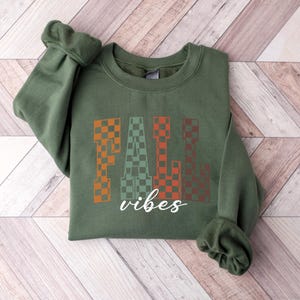 May include: An olive green sweatshirt featuring the text "FALL vibes". The word "FALL" is in a checkered pattern of orange, green, and brown. The word "vibes" is in a white cursive font.