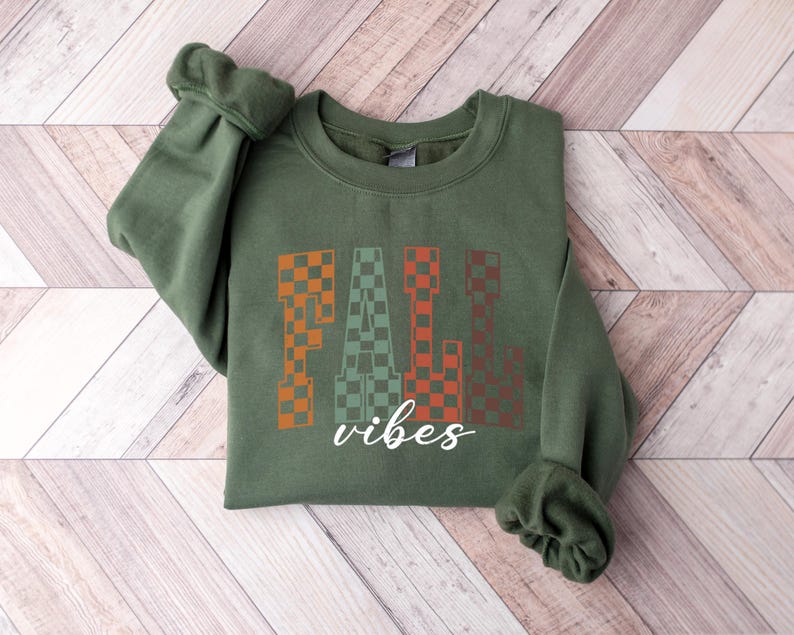 May include: An olive green sweatshirt featuring the text "FALL vibes". The word "FALL" is in a checkered pattern of orange, green, and brown. The word "vibes" is in a white cursive font.