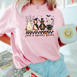 May include: A light pink t-shirt with a Halloween-themed graphic. The graphic features four cartoon geese dressed as a ghost, vampire, pumpkin, and skeleton, with the text "just a spooky goose". A white mug with a yellow smiley face is held in the hand.