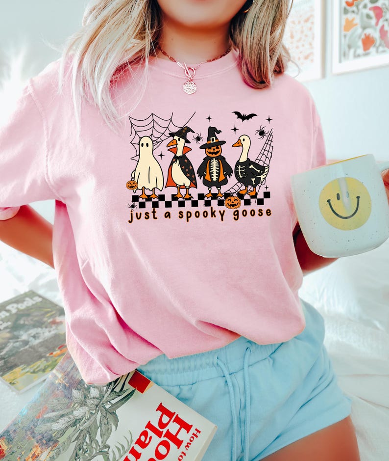 May include: A light pink t-shirt with a Halloween-themed graphic. The graphic features four cartoon geese dressed as a ghost, vampire, pumpkin, and skeleton, with the text "just a spooky goose". A white mug with a yellow smiley face is held in the hand.