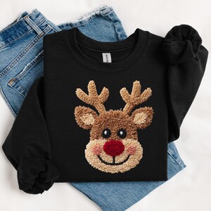 May include: Black sweatshirt featuring a reindeer face with a red nose, brown antlers, and a tan face. The reindeer design is embroidered. The sweatshirt is folded on top of a pair of blue jeans.