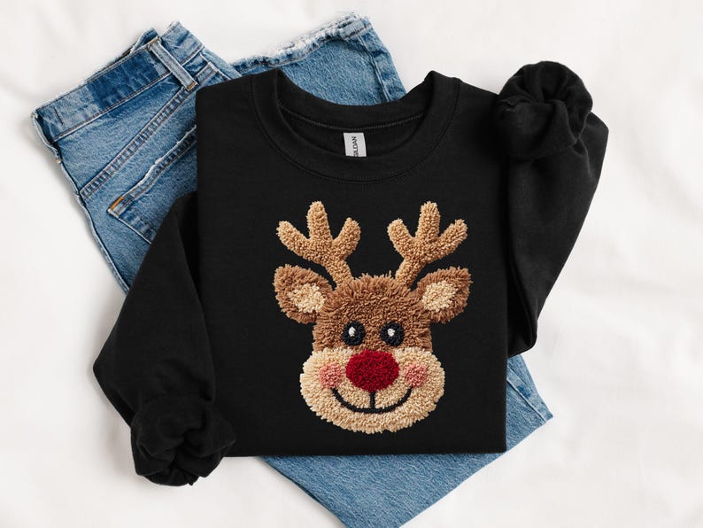May include: Black sweatshirt featuring a reindeer face with a red nose, brown antlers, and a tan face. The reindeer design is embroidered. The sweatshirt is folded on top of a pair of blue jeans.