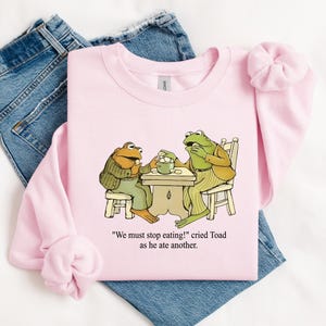 Puede incluir: Sudadera rosa claro con una ilustraci&oacute;n de dibujos animados de dos ranas en una mesa. El texto dice: "&iexcl;Debemos dejar de comer!" grit&oacute; Sapo mientras com&iacute;a otro. La sudadera est&aacute; colocada sobre unos vaqueros azules.