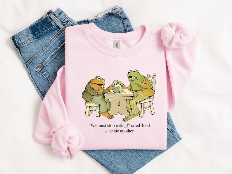 Puede incluir: Sudadera rosa claro con una ilustraci&oacute;n de dibujos animados de dos ranas en una mesa. El texto dice: "&iexcl;Debemos dejar de comer!" grit&oacute; Sapo mientras com&iacute;a otro. La sudadera est&aacute; colocada sobre unos vaqueros azules.