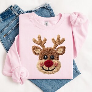 May include: A light pink sweatshirt featuring a reindeer face with brown antlers, a red nose, and a smiling mouth. The sweatshirt is folded on top of a pair of blue jeans. A matching pink scrunchie is on the sleeve.