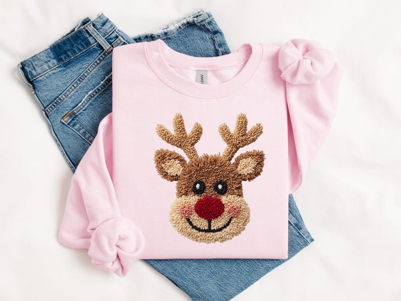 May include: A light pink sweatshirt featuring a reindeer face with brown antlers, a red nose, and a smiling mouth. The sweatshirt is folded on top of a pair of blue jeans. A matching pink scrunchie is on the sleeve.