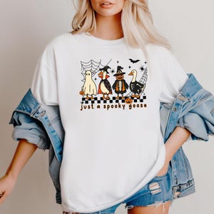 May include: White t-shirt featuring a Halloween-themed graphic. The design includes a ghost, a penguin, a scarecrow, and a goose, all in costumes, with the text "just a spooky goose". The design is set against a black and white checkered pattern.