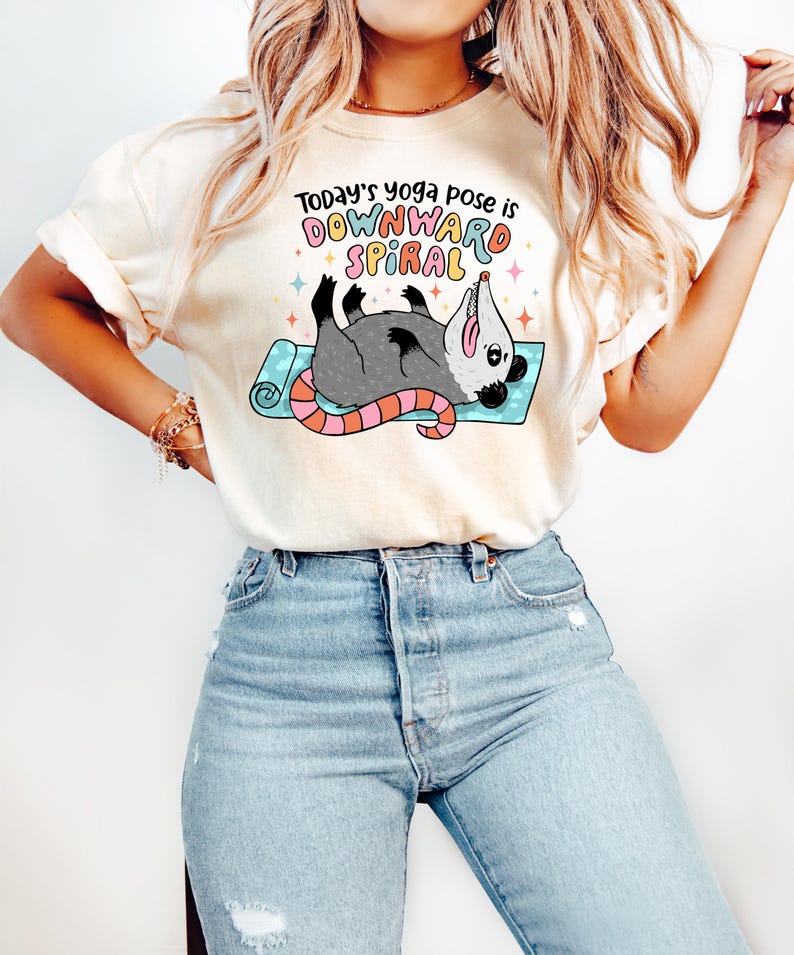 May include: Cream-colored t-shirt with the text "Today's yoga pose is DOWNWARD SPIRAL" and a cartoon opossum on a yoga mat. The opossum is gray with a pink and orange tail. The shirt has a relaxed fit.