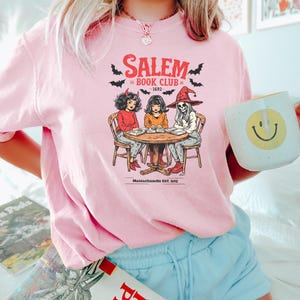 Puede incluir: Camiseta rosa claro con un dise&ntilde;o de "Salem Book Club - 1692" y dibujos de tres figuras alrededor de una mesa. El dise&ntilde;o incluye murci&eacute;lagos y la frase "Massachusetts EST. 1692". La camiseta la lleva una persona.
