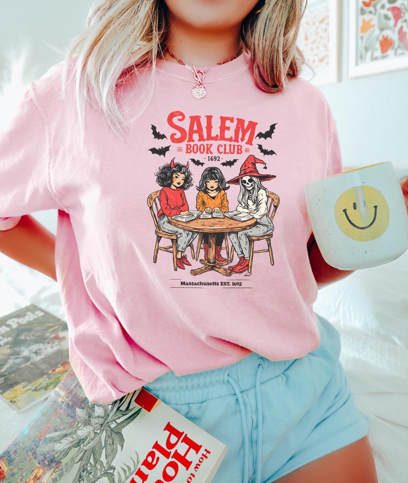Puede incluir: Camiseta rosa claro con un dise&ntilde;o de "Salem Book Club - 1692" y dibujos de tres figuras alrededor de una mesa. El dise&ntilde;o incluye murci&eacute;lagos y la frase "Massachusetts EST. 1692". La camiseta la lleva una persona.