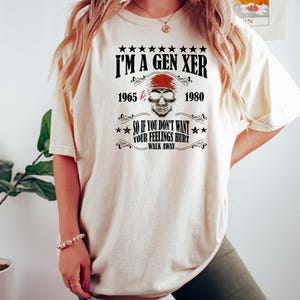 Op de afbeelding: Een wit T-shirt met een zwarte grafische print die "I'm a Gen Xer" zegt met een schedel en gekruiste beenderen en de jaren 1965 en 1980. De tekst onder de schedel zegt "So if you don't want your feelings hurt walk away."