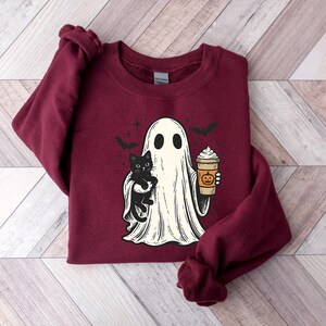 May include: Burgundy sweatshirt with a Halloween graphic. The design shows a ghost holding a black cat and a pumpkin spice latte. Two bats fly above the ghost. The sweatshirt is made of a soft material, ideal for autumn wear.
