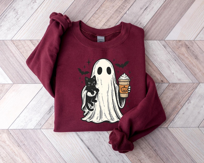 May include: Burgundy sweatshirt with a Halloween graphic. The design shows a ghost holding a black cat and a pumpkin spice latte. Two bats fly above the ghost. The sweatshirt is made of a soft material, ideal for autumn wear.