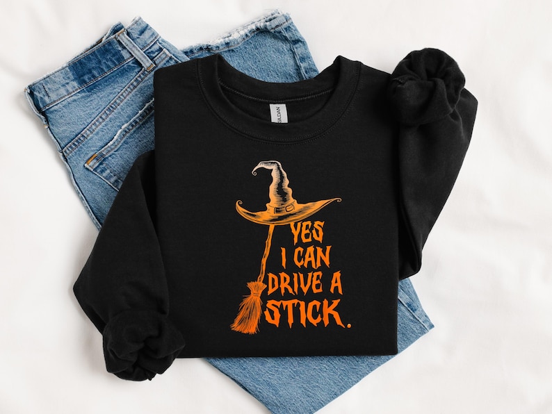 May include: Black crewneck sweatshirt with an orange witch hat and broom graphic. The text "YES I CAN DRIVE A STICK" is printed in orange. The sweatshirt is folded on top of blue jeans.