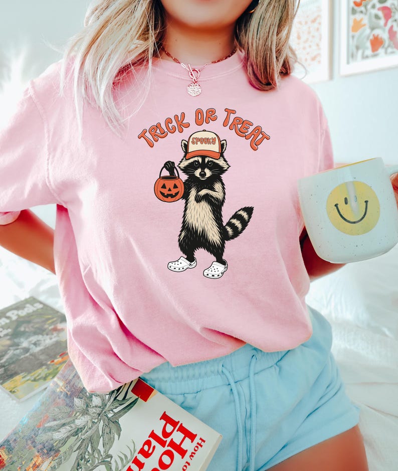 May include: Pink t-shirt with a Halloween-themed graphic. The design features a raccoon wearing a hat that says "Spooky" and holding a pumpkin bucket. The text "Trick or Treat" is arched above the raccoon.