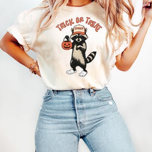 May include: Cream-colored t-shirt featuring a raccoon graphic holding a jack-o'-lantern and wearing a hat. The text "Trick or Treat" is arched above the raccoon. The raccoon is wearing white shoes. The shirt is paired with light blue jeans.