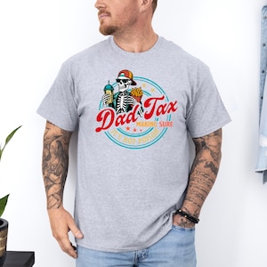 Pode incluir: T-shirt cinza claro com um gr&aacute;fico colorido. O design inclui o texto "Dad Tax" em vermelho, um esqueleto usando um bon&eacute; e a frase "Making Sure It's Not Poison" em um c&iacute;rculo. O esqueleto est&aacute; segurando uma bebida e batatas fritas.