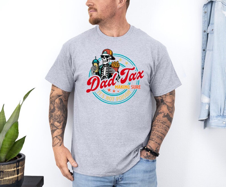 Pode incluir: T-shirt cinza claro com um gr&aacute;fico colorido. O design inclui o texto "Dad Tax" em vermelho, um esqueleto usando um bon&eacute; e a frase "Making Sure It's Not Poison" em um c&iacute;rculo. O esqueleto est&aacute; segurando uma bebida e batatas fritas.