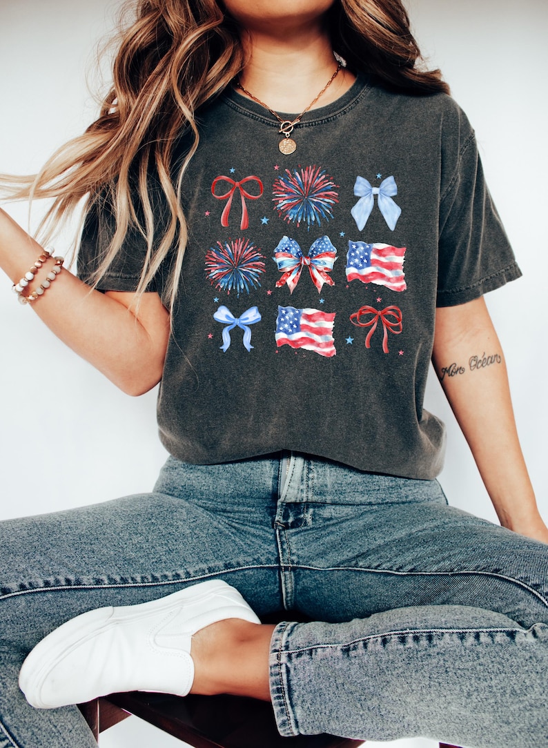 May include: A dark gray t-shirt with a graphic print of red, white, and blue bows, fireworks, and American flags.