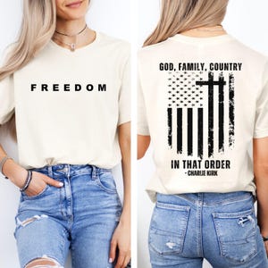 May include: Cream-colored t-shirt with the word "FREEDOM" printed in black on the front. The back of the shirt features a distressed American flag design with a cross and the text "GOD, FAMILY, COUNTRY IN THAT ORDER - CHARLIE KIRK."
