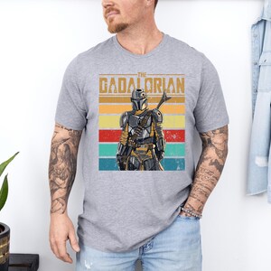 K&ouml;nnte beinhalten: Graues T-Shirt mit einem Retro-Grafikdesign eines Charakters im Mandalorianer-Stil und dem Text "The Gadalorian". Das Design zeigt einen Farbblock aus gelben, orangefarbenen, roten und t&uuml;rkisfarbenen Streifen. Das T-Shirt hat einen Rundhalsausschnitt.