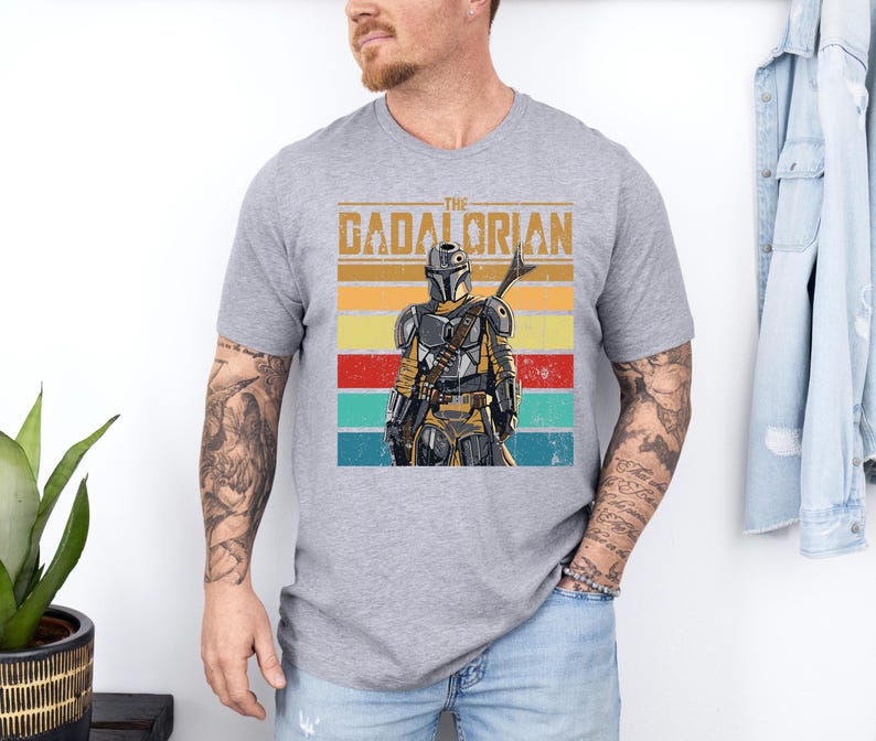 K&ouml;nnte beinhalten: Graues T-Shirt mit einem Retro-Grafikdesign eines Charakters im Mandalorianer-Stil und dem Text "The Gadalorian". Das Design zeigt einen Farbblock aus gelben, orangefarbenen, roten und t&uuml;rkisfarbenen Streifen. Das T-Shirt hat einen Rundhalsausschnitt.