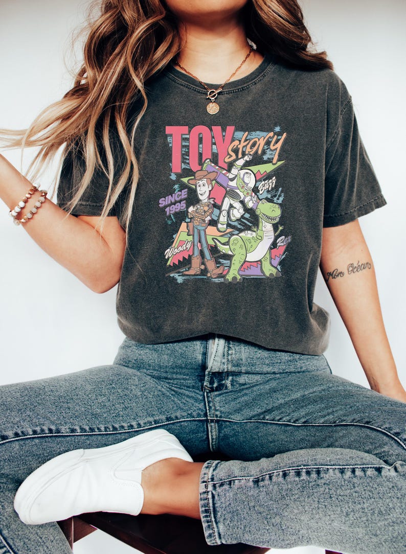 Puede incluir: Camiseta gris oscuro con un gr&aacute;fico colorido de los personajes de Toy Story, incluyendo a Woody, Buzz Lightyear y Rex. La camiseta tiene el texto "TOY STORY" y "SINCE 1995". La camiseta se combina con vaqueros azules y zapatos blancos.