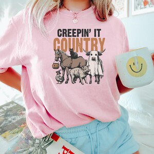 May include: Pink t-shirt with the phrase "CREEPIN' IT COUNTRY" in black and brown. The graphic features a horse, cat, cow, pig, and chicken, all in Halloween costumes. The person is holding a white mug with a yellow smiley face.