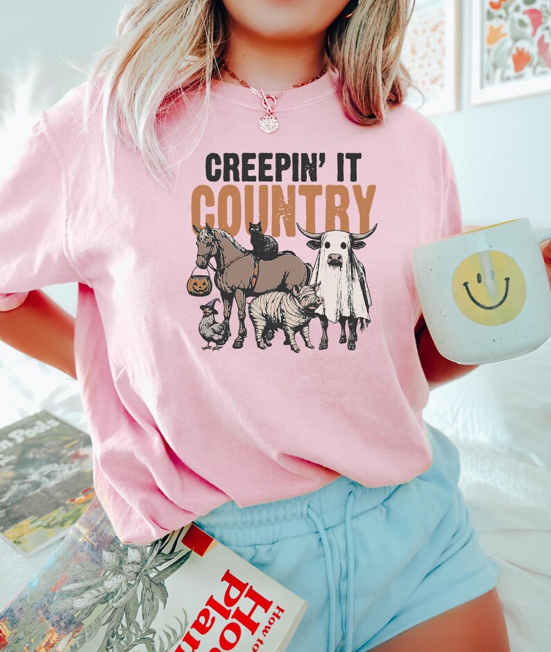 May include: Pink t-shirt with the phrase "CREEPIN' IT COUNTRY" in black and brown. The graphic features a horse, cat, cow, pig, and chicken, all in Halloween costumes. The person is holding a white mug with a yellow smiley face.