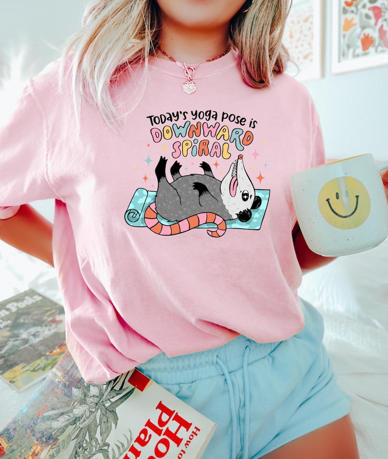 May include: Pink sweatshirt with the text "Today's yoga pose is Downward Spiral" and a cartoon opossum on a yoga mat. The opossum is gray with a pink nose and tail. A white mug with a yellow smiley face is held in the hand.