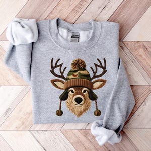 Puede incluir: Sudadera gris claro con un dise&ntilde;o de cara de ciervo con un gorro marr&oacute;n y camuflaje. El ciervo tiene cuernos. La sudadera est&aacute; colocada sobre una superficie de madera.