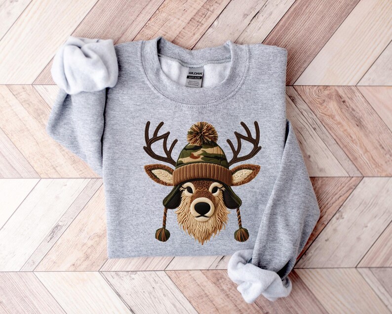 Puede incluir: Sudadera gris claro con un dise&ntilde;o de cara de ciervo con un gorro marr&oacute;n y camuflaje. El ciervo tiene cuernos. La sudadera est&aacute; colocada sobre una superficie de madera.