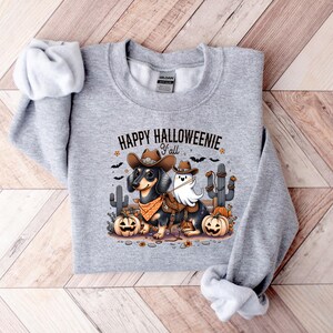 May include: A light grey sweatshirt with the text "HAPPY HALLOWEENIE Y'all". The graphic features two dachshunds in Halloween costumes, one as a cowboy and the other as a ghost, with pumpkins and cacti.