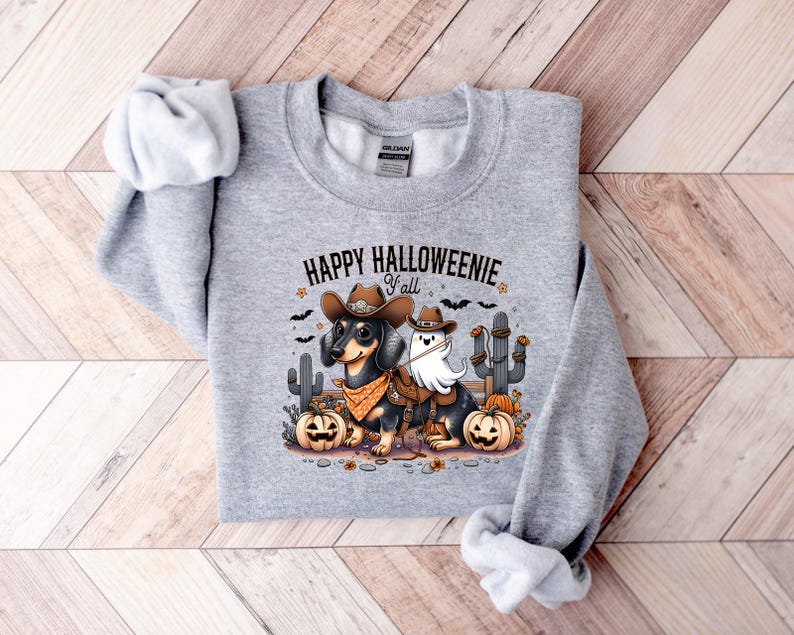 May include: A light grey sweatshirt with the text "HAPPY HALLOWEENIE Y'all". The graphic features two dachshunds in Halloween costumes, one as a cowboy and the other as a ghost, with pumpkins and cacti.