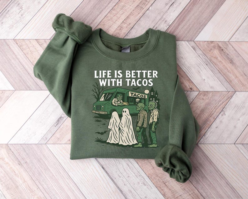 May include: Green sweatshirt with the phrase "LIFE IS BETTER WITH TACOS" and a graphic of a taco truck, zombies, and ghosts. The design is in white, green, and black. The sweatshirt is made of a soft material.